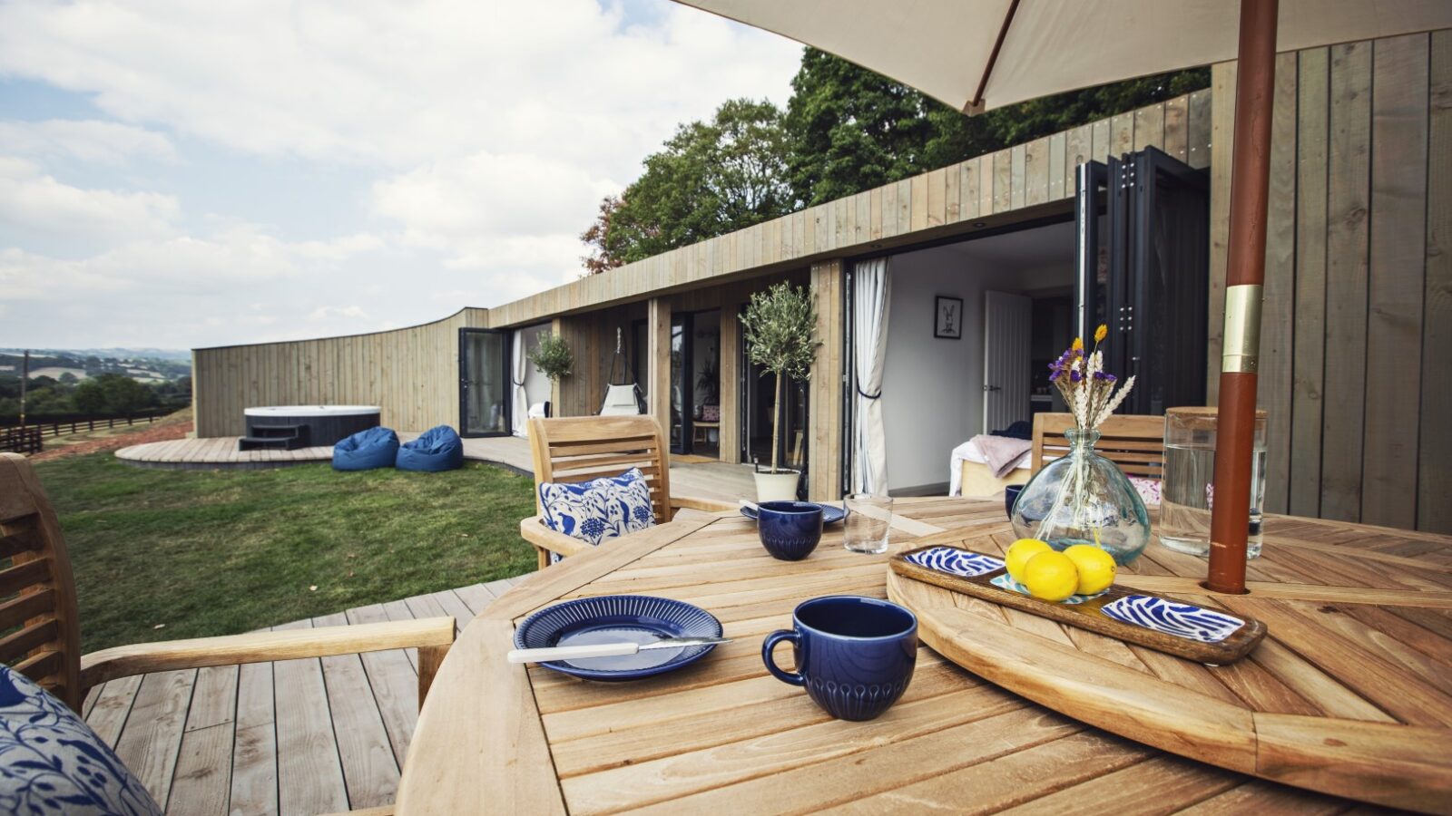 On the wooden deck, a table is set for breakfast with plates and cups, offering a serene escape at Pontypinna's hideaways. In the background, an elegant curved wooden building complements the cozy outdoor seating.