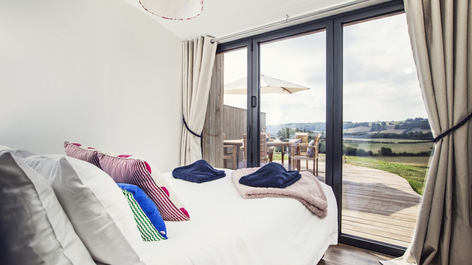 This serene bedroom at Pontypinna features a white bed adorned with colorful pillows and neatly folded towels. Large glass doors lead to a deck, offering outdoor seating and a scenic view, perfect for peaceful hideaways.