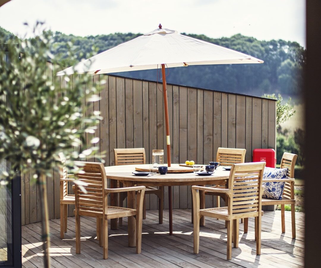 The outdoor patio at Pontypinna Hideaways features a charming wooden dining set, umbrella, and lush plants, all surrounded by a wooden fence that opens up to breathtaking views of the green hills.