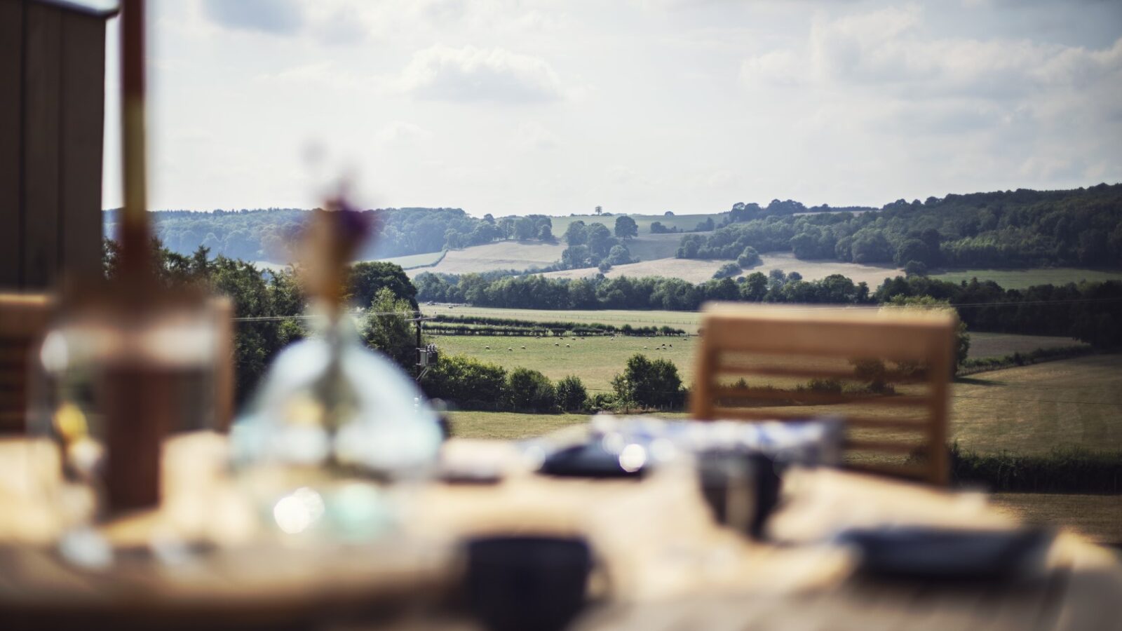 A blurred view of a table and chairs on a patio at Pontypinna Hideaways overlooks a rural landscape with fields and trees under a cloudy sky.