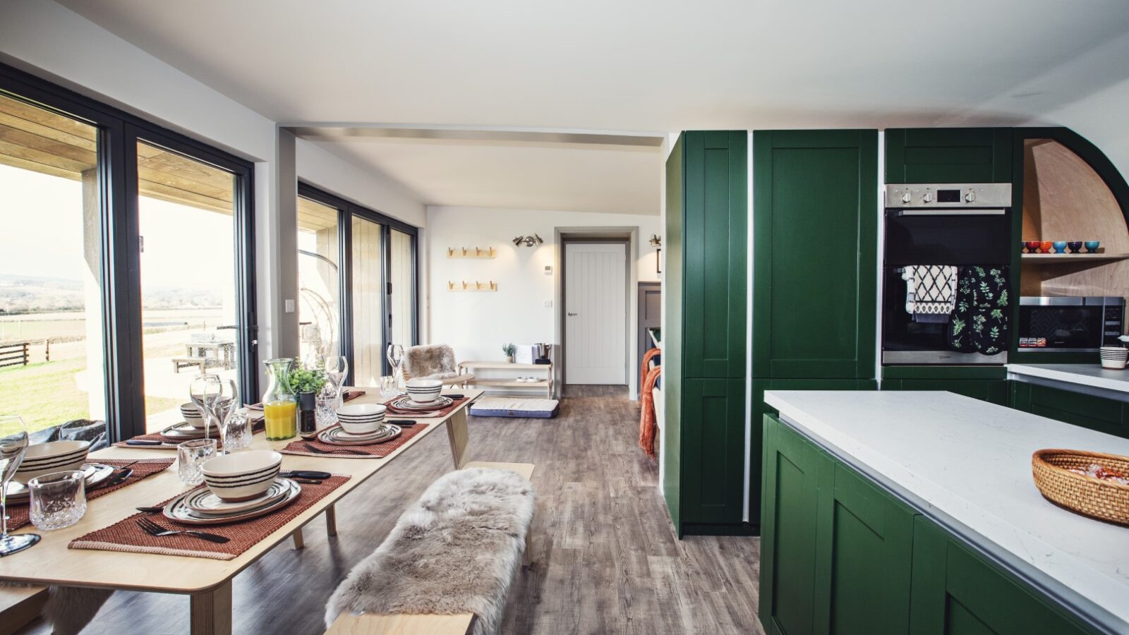 Discover the modern kitchen and dining area at Pontypinna Hideaways, featuring elegant green cabinets, a long table set with dishes, and large windows offering a scenic view over tranquil hideaways.