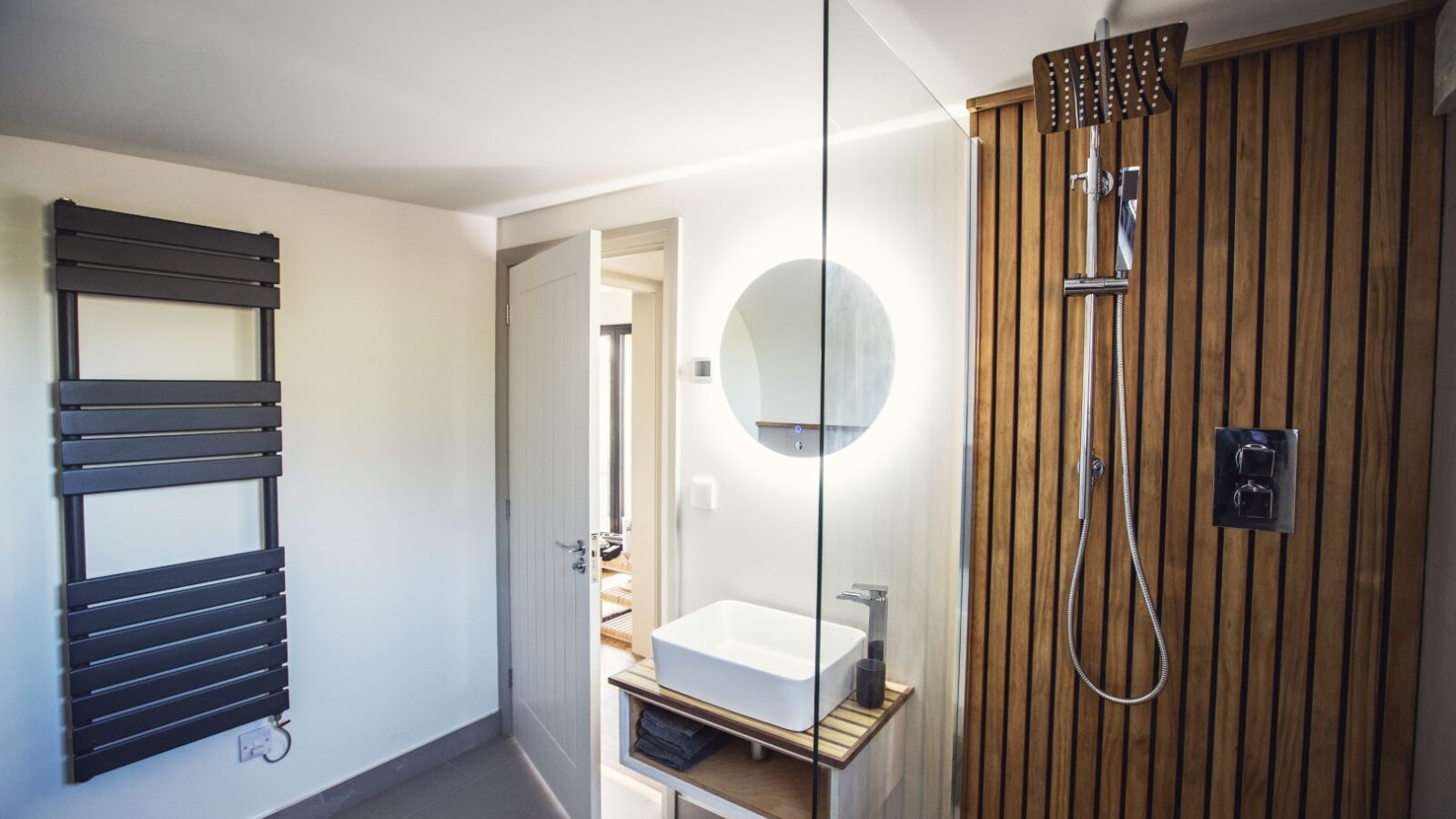 Discover a modern bathroom hideaway featuring a glass shower, round mirror above the sink, and elegant wooden wall paneling. Experience warmth with a wall-mounted towel warmer in this Pontypinna-inspired design.