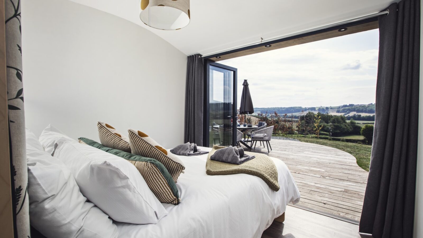 The bedroom at Pontypinna Hideaways features a neatly made bed overlooking a wooden deck and outdoor seating area, with a scenic view in the background.