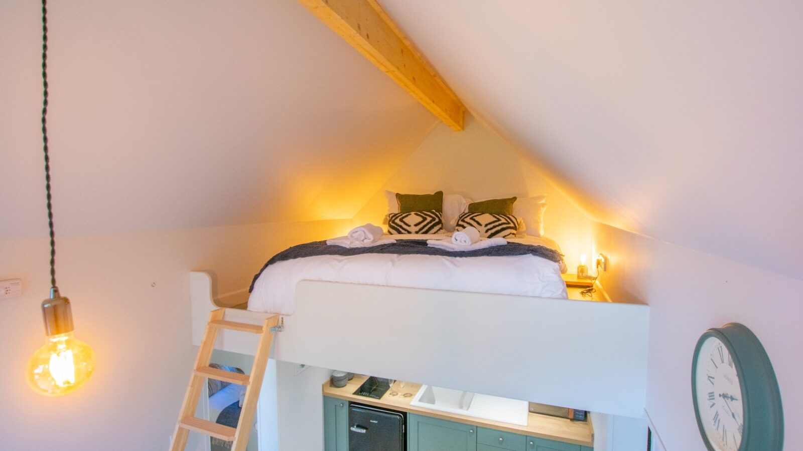 A cozy loft bedroom with a wooden beam ceiling features a double bed adorned with pillows and rolled towels, perfect for glamping enthusiasts. Below, there's a kitchenette with green cabinets. A vintage-style wall clock and hanging light bulb enhance the quaint atmosphere of this Porth Eryri retreat.