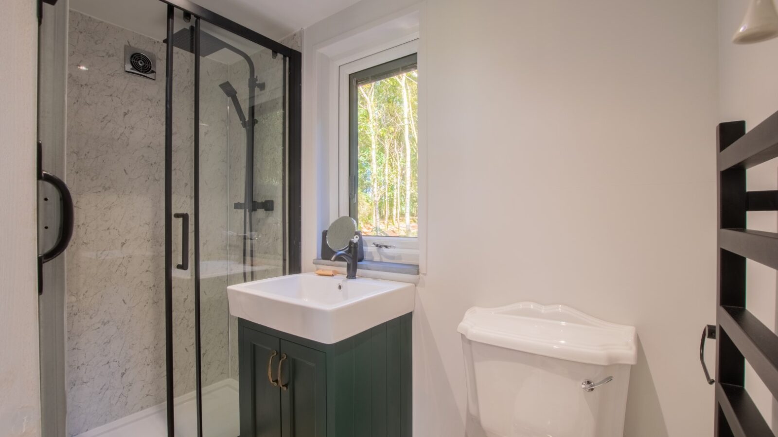 A modern bathroom at Porth Eryri Glamping features a glass-enclosed shower with a black frame, a white sink set on a dark green vanity, and a white toilet. A small window offers views of trees above the sink, while the light gray walls add to the serene glamping experience.