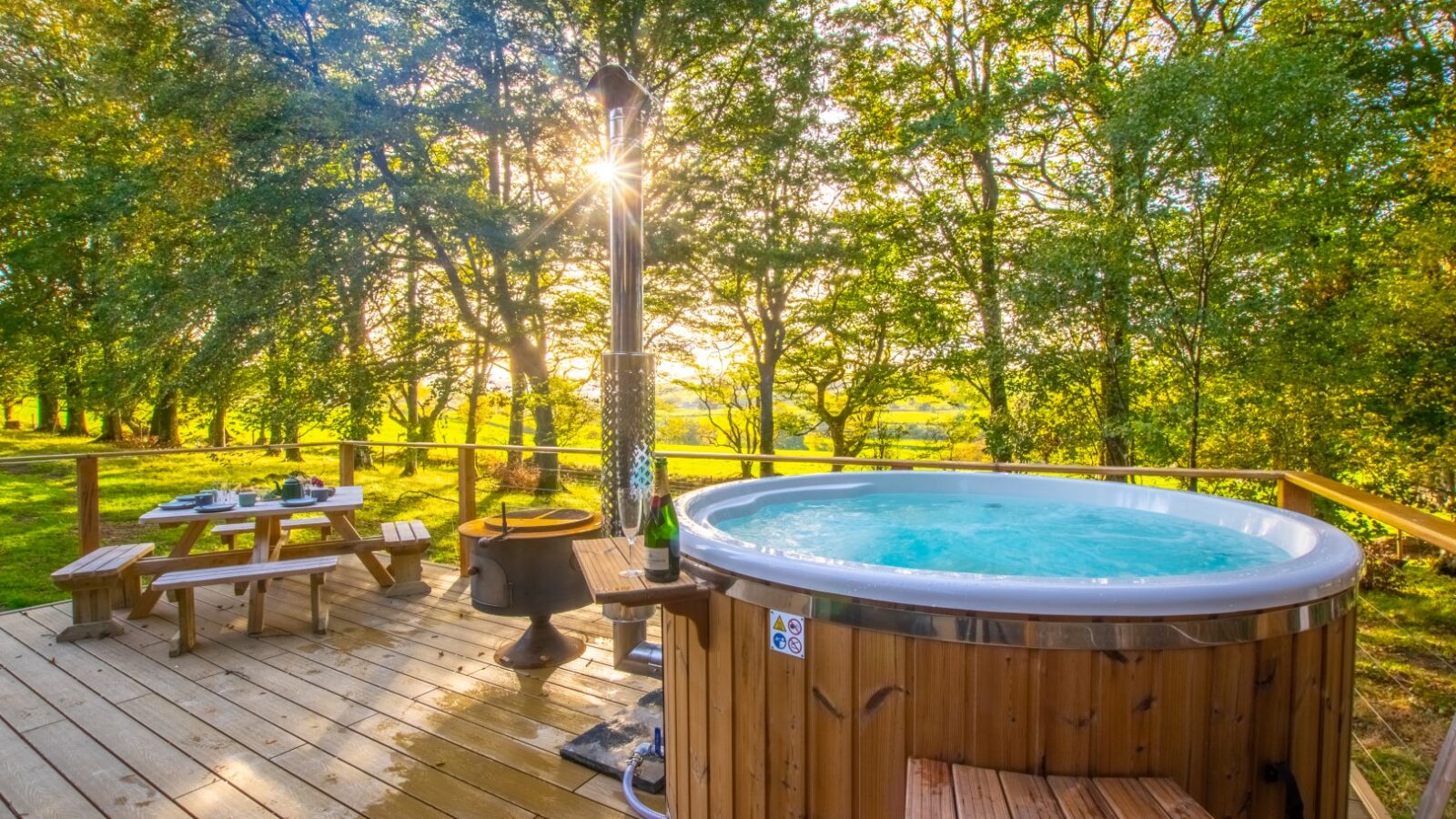 A serene outdoor deck at Porth Eryri Glamping features a bubbling hot tub, a cozy fire pit, and a picnic table set with dishes. It's surrounded by lush green trees under a bright, sunny sky.