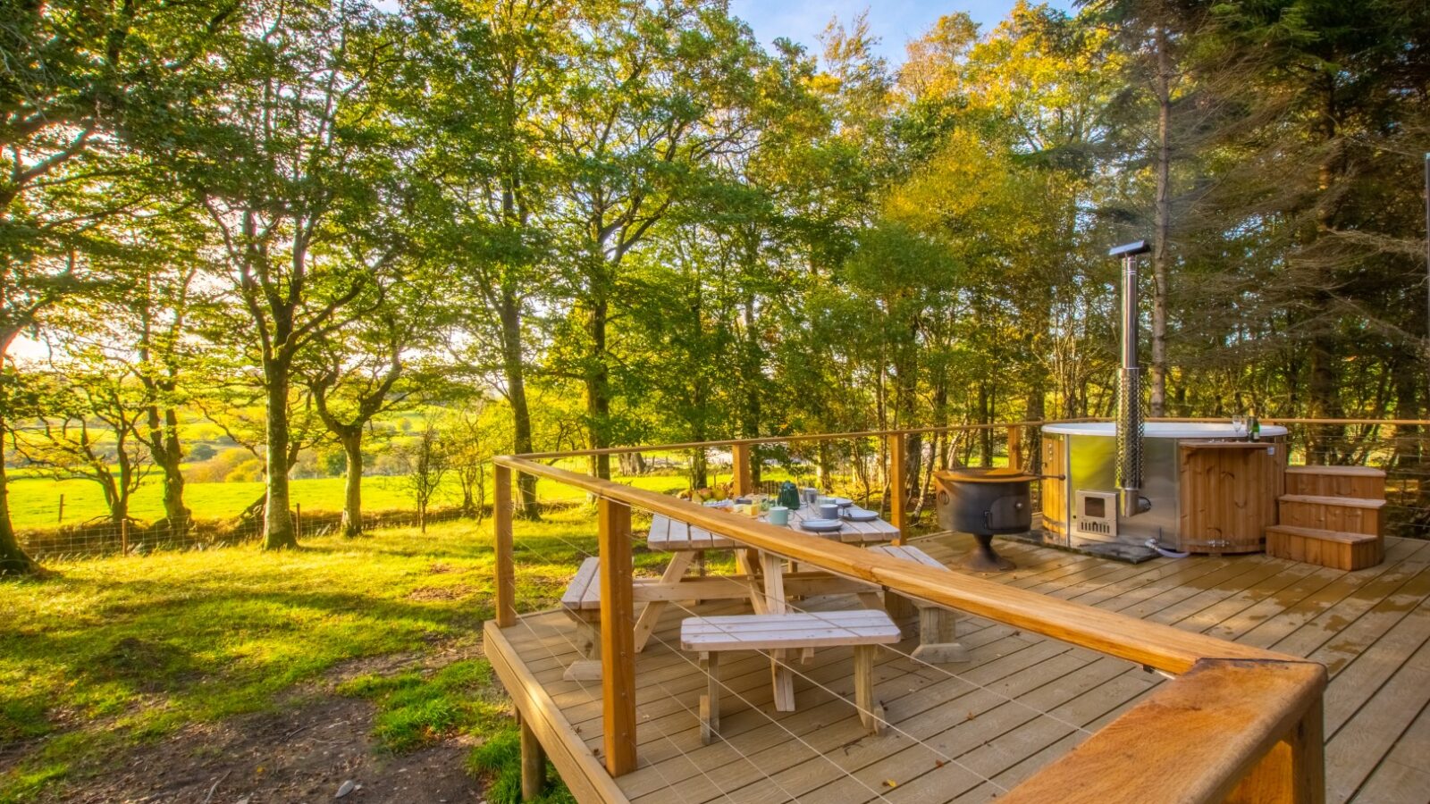 A scenic outdoor deck, perfect for glamping, is surrounded by lush green trees under a clear sky. The deck features a wooden hot tub, a small stove, and a picnic table with benches. Sunlight filters through the branches, casting a warm glow across this enchanting haven near Porth Eryri.