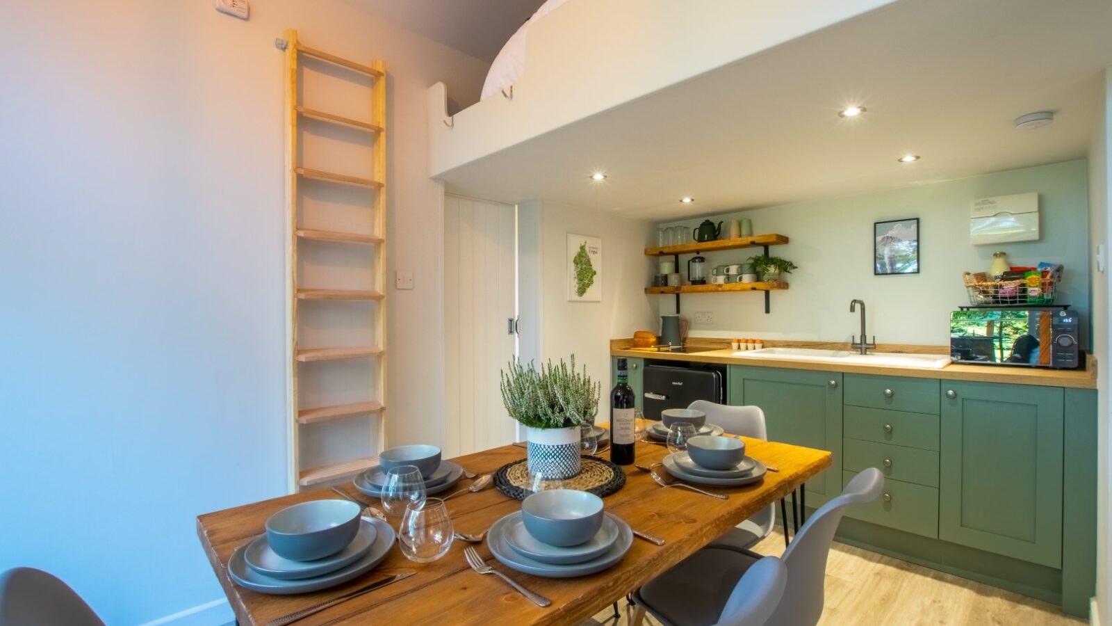 A compact kitchen and dining area, perfect for glamping at Porth Eryri, features a wooden table set with gray dishes and utensils. A green kitchenette with open shelves stands in the background, while a wooden ladder leads to a lofted bed above. The space is well-lit and modern.