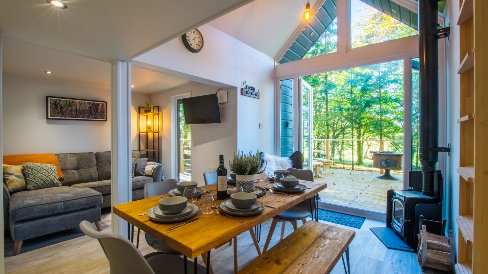 A cozy cabin interior exudes glamping charm with a wooden dining table set for four, a gray sofa, and a wall-mounted TV. Large glass doors open to a deck showcasing Porth Eryri's stunning trees and sunshine. The space is bright and modern, with a warm, inviting atmosphere.