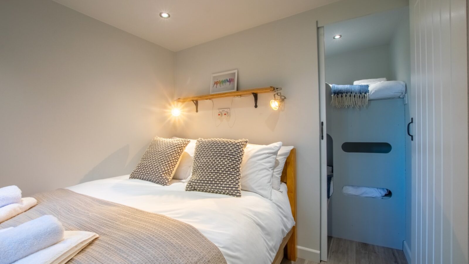 A cozy glamping bedroom in Porth Eryri, featuring a double bed with white bedding and gray pillows. To the right, there's a small room with bunk beds. Warm lighting accents the wooden shelf holding a picture frame above the bed, where towels are neatly placed.