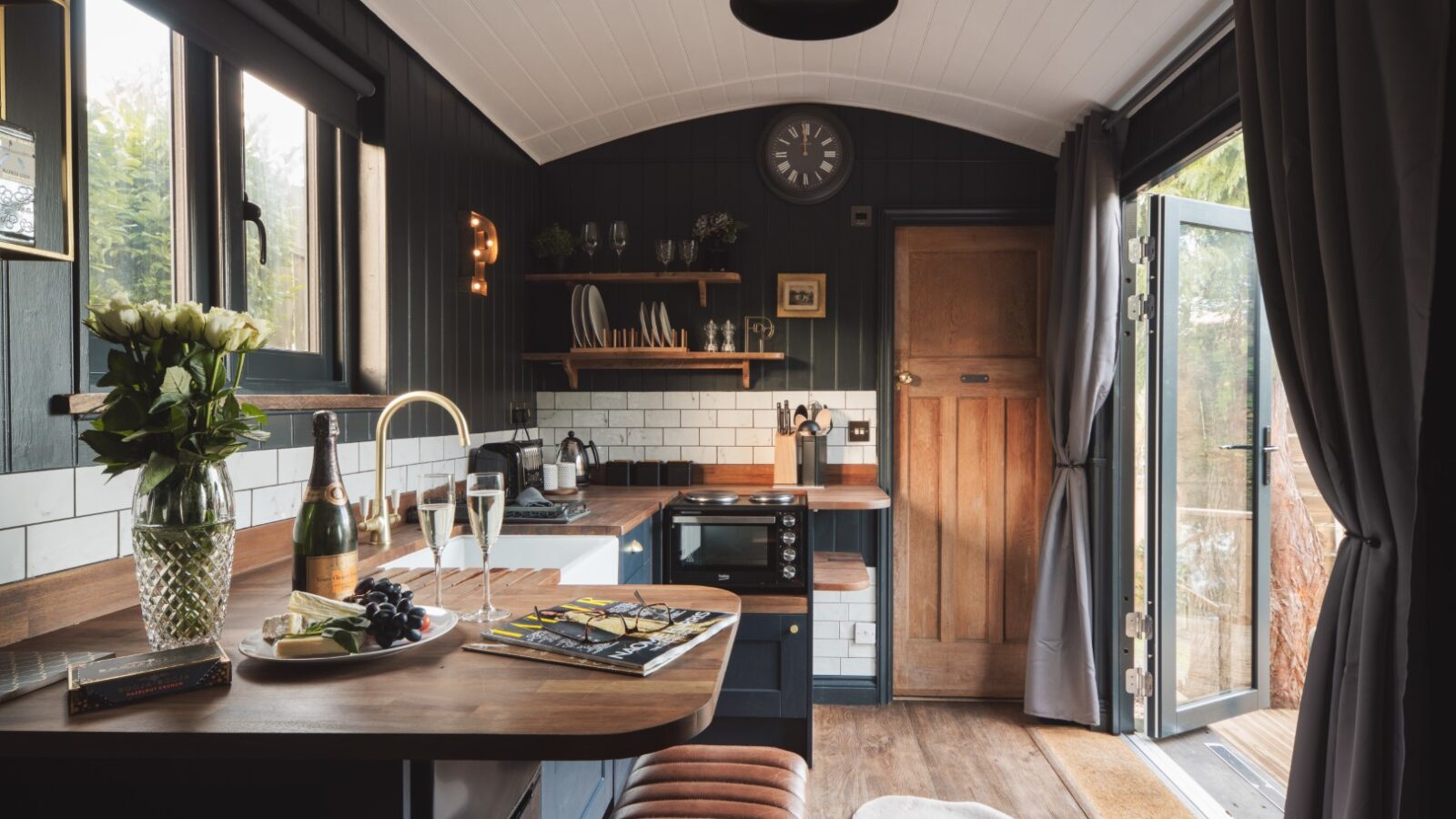 Cozy kitchen in a Shepherds Hut Retreat with a wooden countertop, flowers, wine, and snacks. Open doors lead outside; shelves and clock adorn the walls.