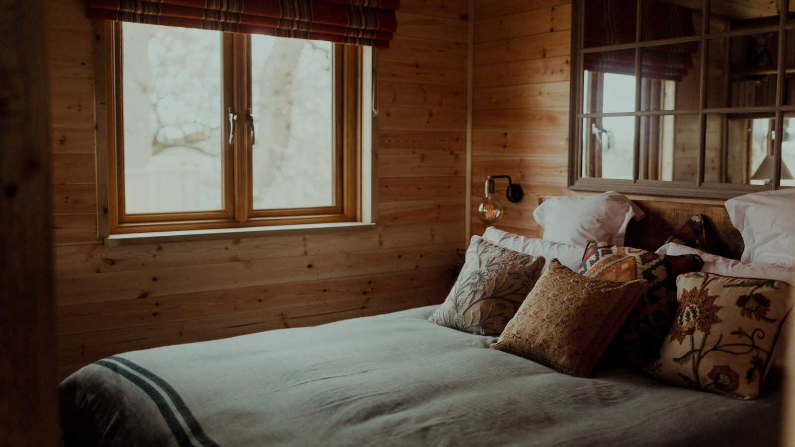 A cozy bedroom with wooden walls and a large window, resembling the serene comfort of a Netherby Treehouse. The bed is made up with a light-colored comforter and several decorative pillows. A striped Roman shade covers the window, and a wall lamp is attached to the headboard. The room has a warm and rustic feel.