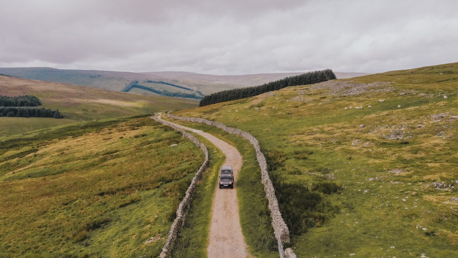 A Defender cruises along a narrow dirt road flanked by stone walls and rolling green hills, perfect for a camping adventure under the cloudy sky.