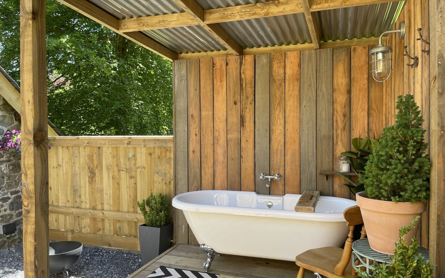 Nestled in the heart of Trallwm's lush forests, this outdoor wooden bathroom features a white freestanding tub, a small wooden chair, potted plants, and a zigzag rug, creating a serene cottage-like atmosphere.