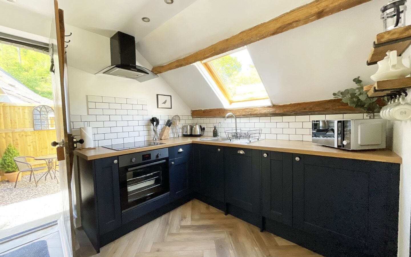 Modern kitchen with dark cabinets, wooden countertops, skylight, herringbone floor, oven, microwave, and an open door to the patio offering a tranquil view reminiscent of the serene Trallwm Forest.