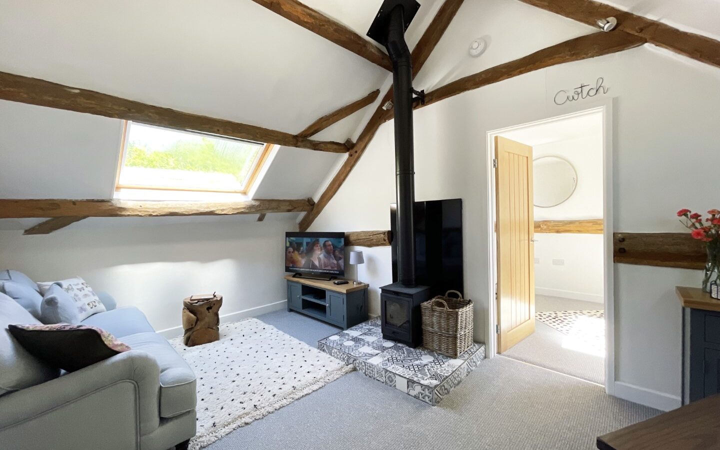 Cozy attic living room with exposed wooden beams, a skylight, wood-burning stove, TV, and sofa. This charming space feels like a Trallwm cottage with its rustic charm and inviting warmth. A small hallway to the right completes this delightful retreat surrounded by the forest's beauty.