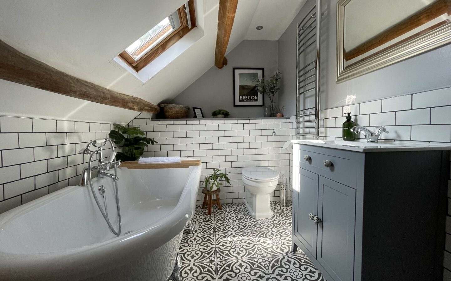 Nestled in the heart of Trallwm Forest, this attic bathroom features a freestanding bathtub, patterned tile floor, skylight, sink with cabinet, and wall-mounted towel rack—offering a perfect retreat within charming forest cottages.