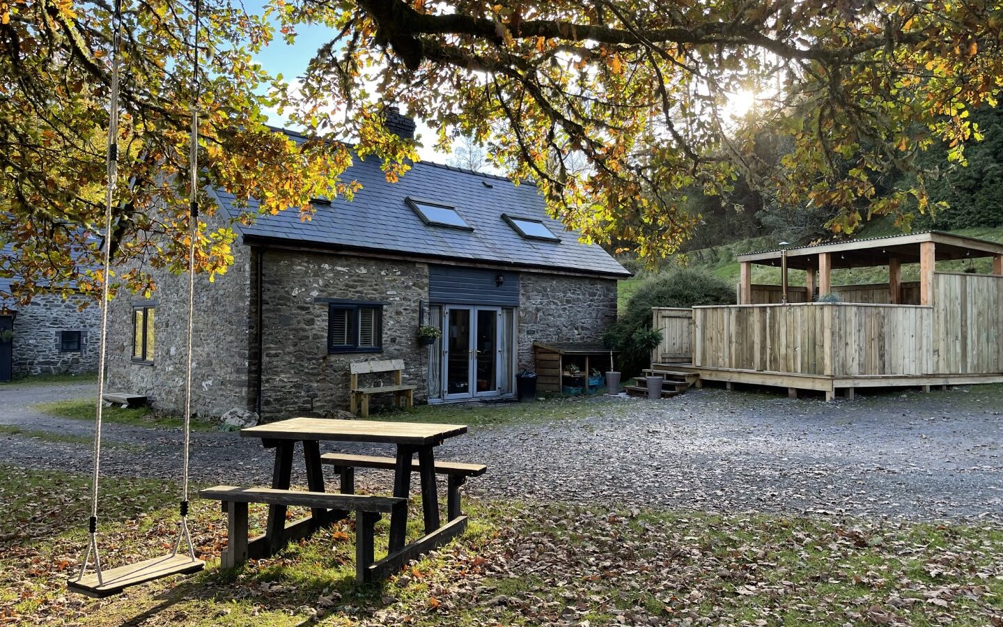 Nestled in the rural tranquility of Trallwm, this stone cottage boasts a wooden deck, picnic table, and a charming rope swing under trees adorned with autumn leaves, perfectly blending with the enchanting forest surroundings.