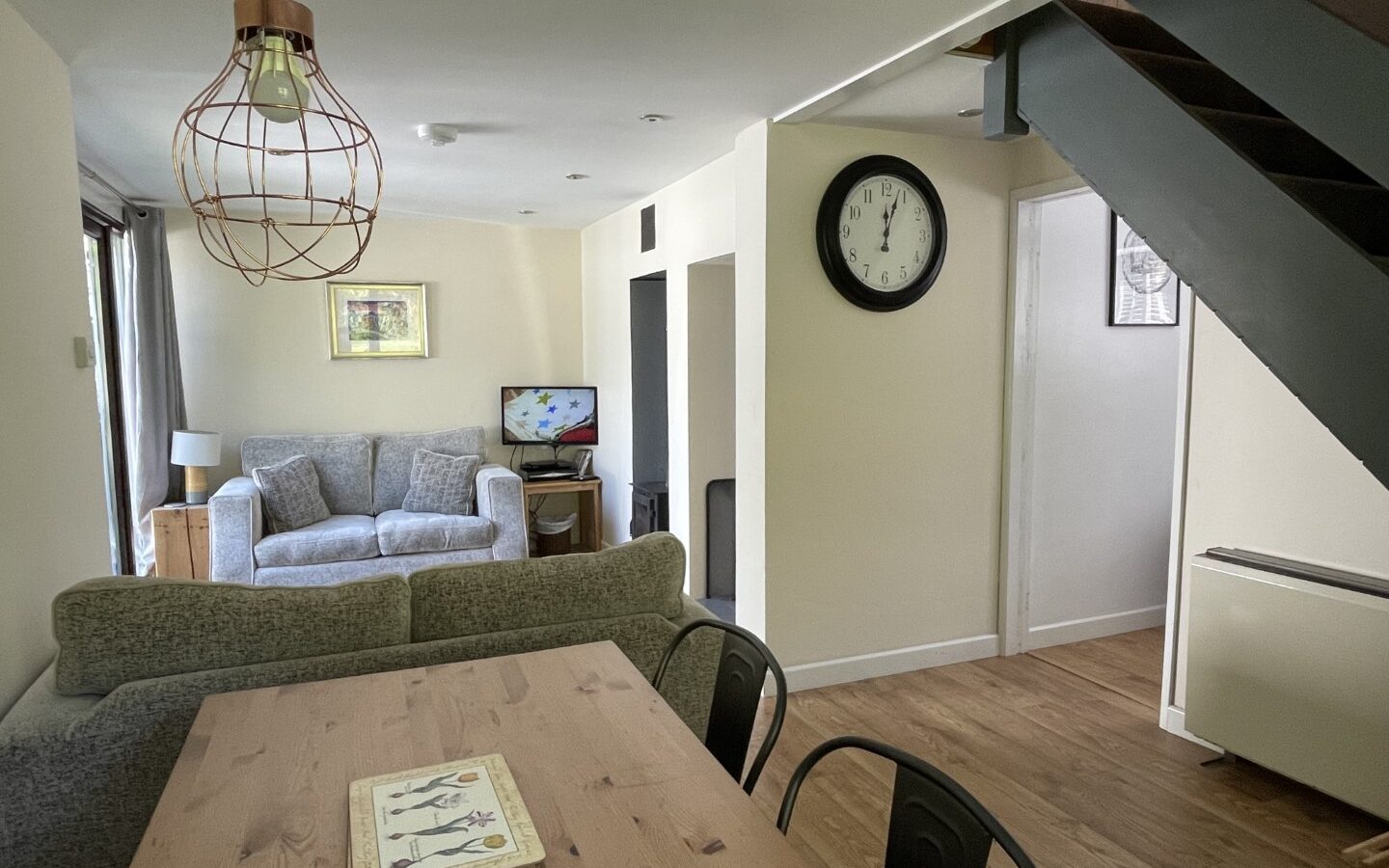 The living room in Trallwm Forest Cottages features a gray sofa, TV, wall clock, and dining area. Wood flooring and a staircase add to its rustic charm.