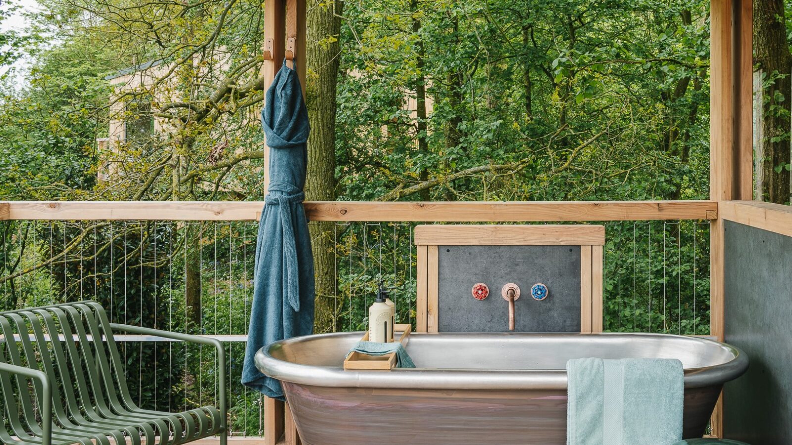 A rustic outdoor area featuring a large metal bathtub surrounded by wooden railing and lush green trees, reminiscent of a treehouse retreat. A metallic chair with green cushions sits nearby, and towels and bath essentials are neatly arranged on and around the tub, creating a serene, Rewild Things spa-like atmosphere.