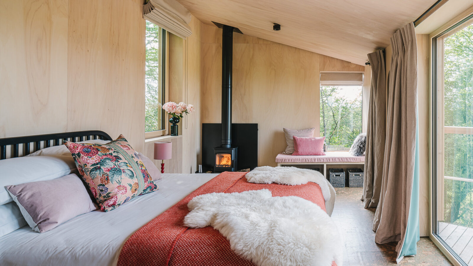 A cozy, light-filled cabin bedroom with light wooden walls and ceiling. The room features a bed dressed in white and red bedding with floral and fur accents. A wood-burning stove stands near the window, and a cushioned seating area by the glass doors leads to a deck overlooking treehouses amidst the forest view.