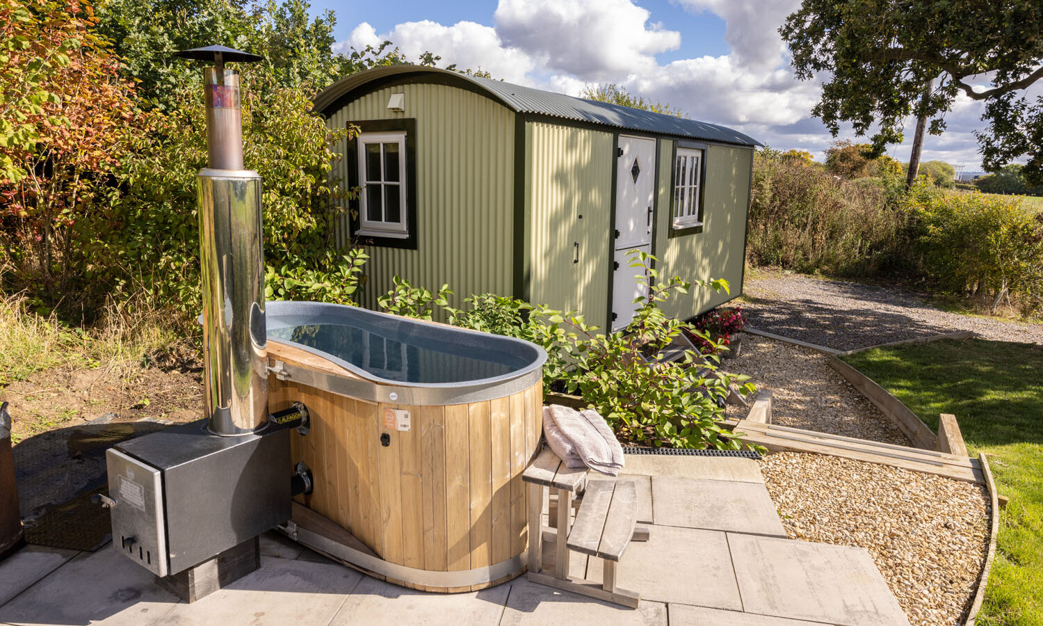 A wooden hot tub with a metal chimney on a stone patio graces the garden at Abbey Farm. Nearby, a green corrugated metal shepherd's hut with a white door and window stands amidst trees under a partly cloudy sky. A wooden chair and towel await beside the tub, inviting relaxation.