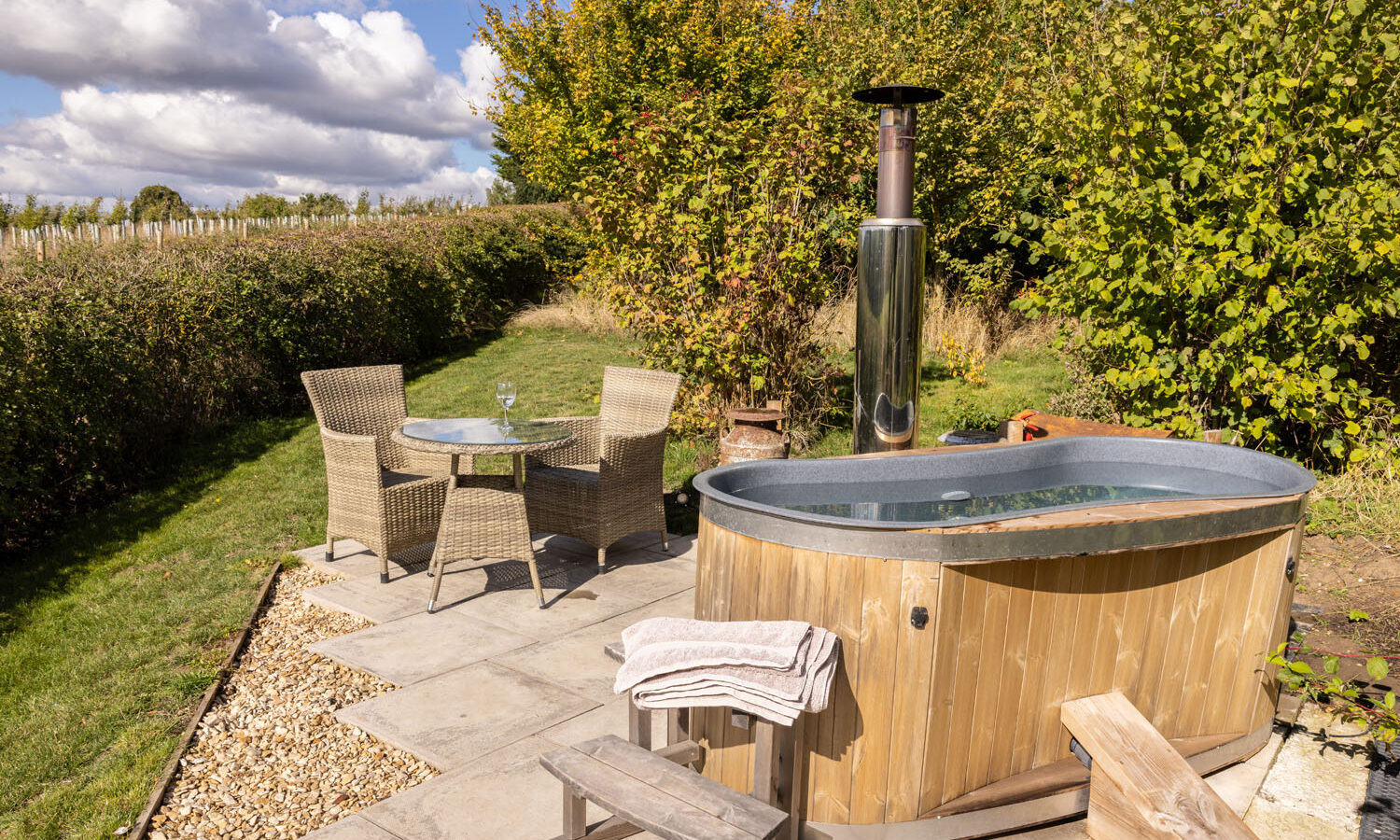 A wooden hot tub is set on a stone patio in the garden of Abbey Farm. Nearby, two wicker chairs and a small glass table with a glass of wine invite relaxation, surrounded by lush green shrubs and trees under a partly cloudy sky.