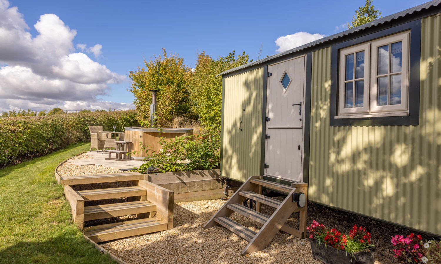 A green cabin reminiscent of charming Shepherds Huts with a white door and two windows sits beside a small wooden deck. The deck has stairs leading down to a gravel path with outdoor furniture and a hot tub. Red flowers bloom in a planter, surrounded by greenery under a blue sky with clouds.