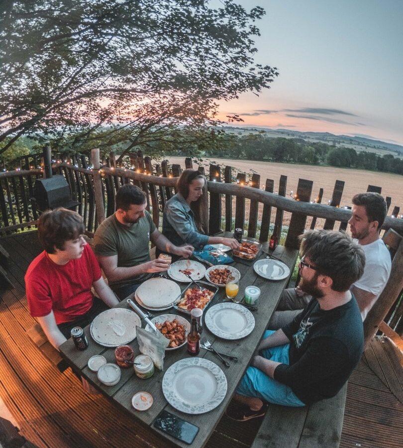 Five people sit around a wooden table outdoors at Baxby Manor, enjoying a meal with various dishes and drinks. The scene is set against a scenic backdrop of trees and a sunset sky. They appear to be engaged in conversation, creating a warm and relaxed atmosphere near Rufus's Roost Treehouse.