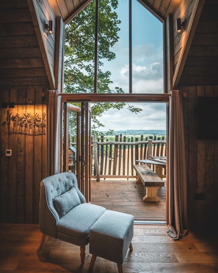 A cozy, rustic room at Rufus's Roost Treehouse with a high vaulted ceiling features a padded lounge chair and ottoman. Large windows offer a view of an outdoor wooden deck with a picnic table, beyond which lies a scenic landscape of green fields and a partly cloudy sky.