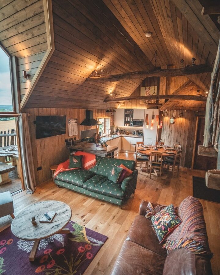 A cozy wooden cabin interior with a vaulted ceiling at Rufus's Roost Treehouse. It features a green sofa, a leather couch, a round coffee table, and a patterned rug. The dining area has wooden table and chairs, and the kitchen is equipped with modern amenities. Warm lighting enhances the space at Baxby Manor.