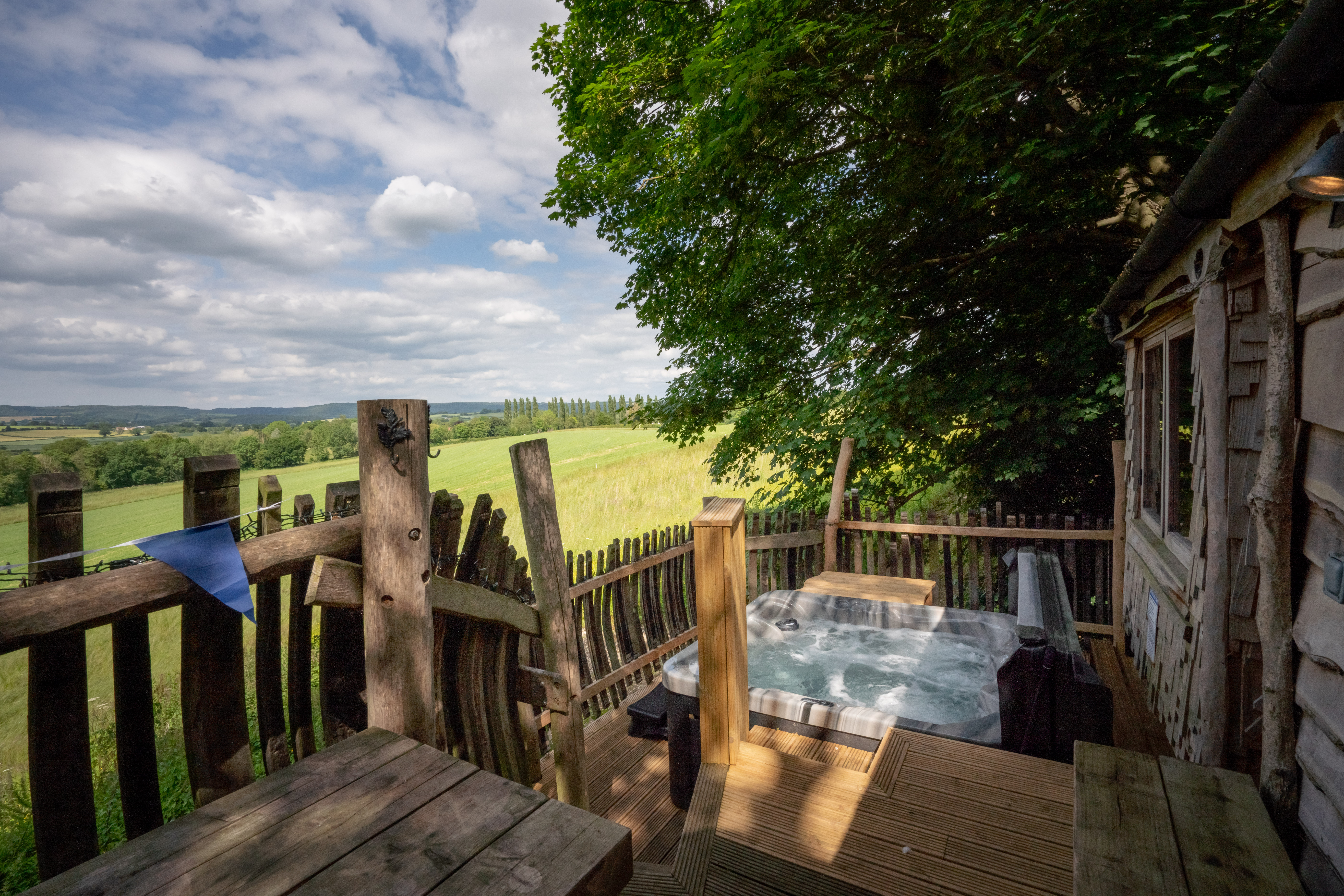 A rustic wooden deck with a hot tub overlooks the scenic countryside of Baxby Manor. The deck, surrounded by a wooden fence and shaded by trees, offers views of vast green fields under a partly cloudy sky. In the foreground sits a wooden table, adding charm to this idyllic setting.