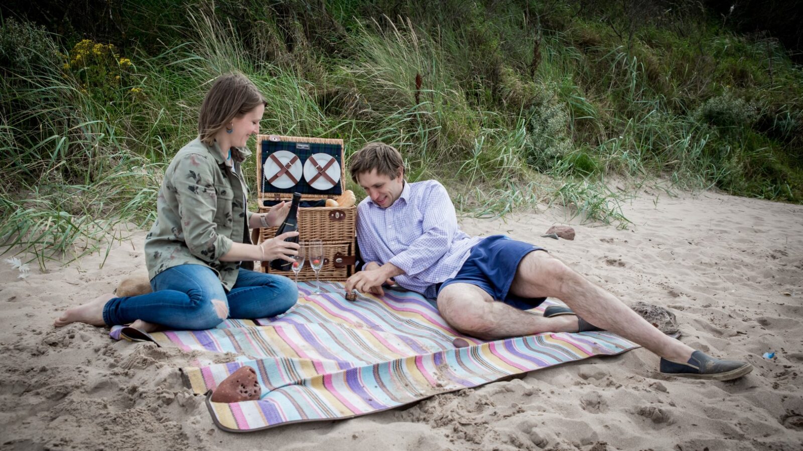 A woman and man sit on a striped blanket on a sandy beach, setting up an old-fashioned radio between them as the waves softly crash against the rocks.