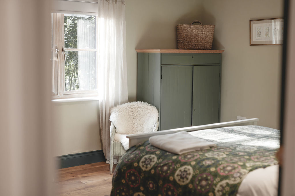 Discover a cozy bedroom designed for secret escapes, featuring a wooden bed with a floral bedspread, a green wardrobe crafted by Browning Bros, a wicker chair, and a basket on top. Natural light filters through the window with charming curtains.