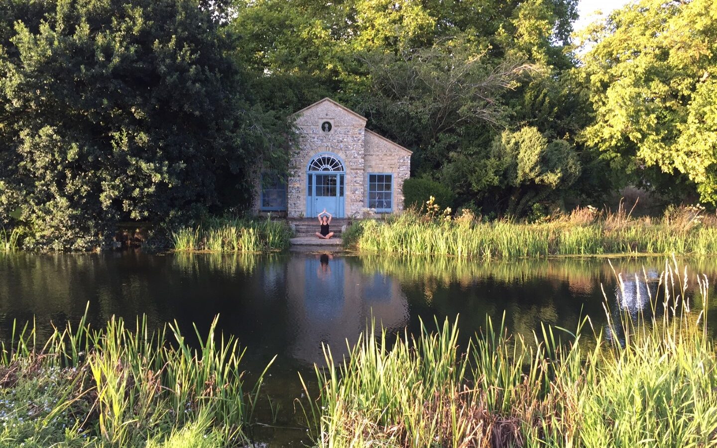 A small stone building with blue windows sits across a pond surrounded by greenery and tall grass, reminiscent of the serene charm found at West Lexham Cabins.