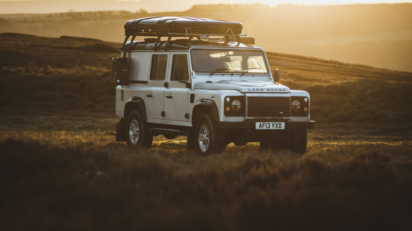 A white Defender off-road vehicle with a roof rack is parked in a grassy field during sunset, perfect for camping adventures, with hills gently rising in the background.