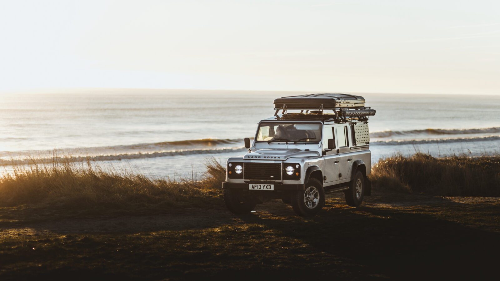 A white Defender with roof cargo drives along the beach at sunset, perfectly capturing the spirit of camping adventure.