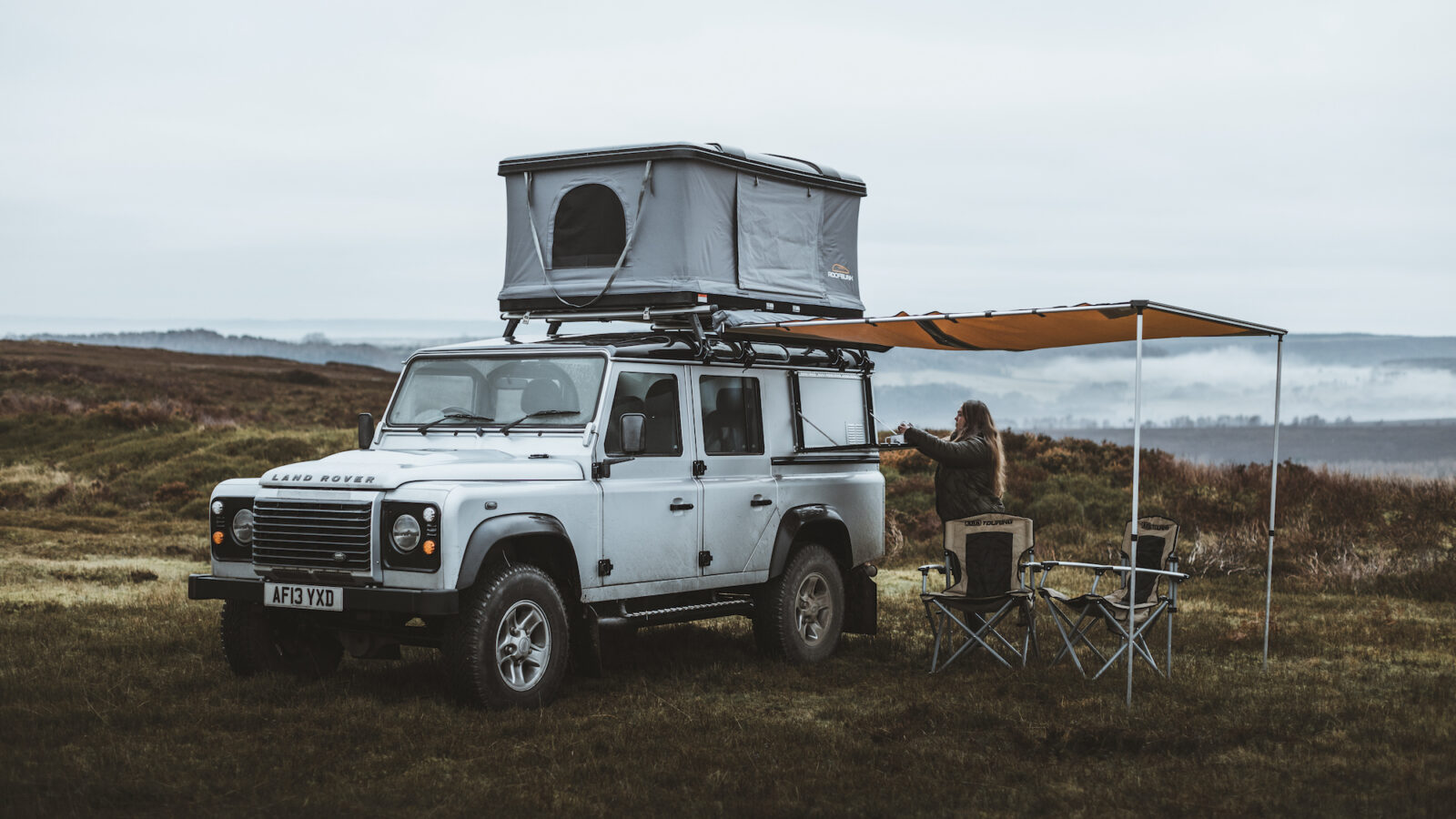 A silver Defender SUV with a roof tent and awning is parked on a grassy field, capturing the essence of camping. A person stands nearby while two chairs are set up, inviting a perfect retreat into nature's embrace.