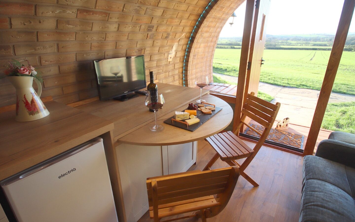 Cozy room with a small table and chairs, TV, mini fridge, wine, and snacks. Open doors offer a view of the grassy Yorkshire landscape reminiscent of charming burrows in the countryside.