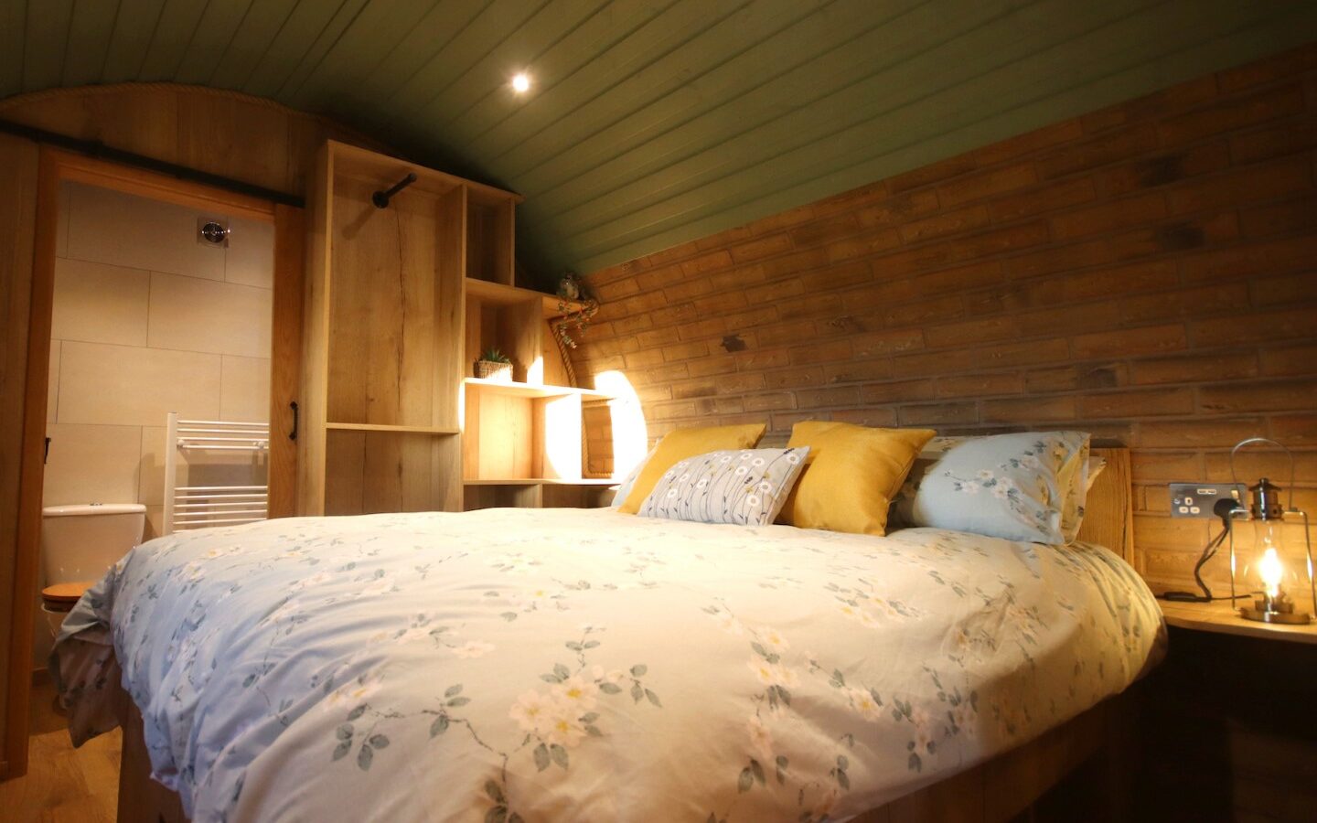 A cozy Yorkshire bedroom with a floral bedspread, warm lighting, and wooden and brick accents exudes the charm of a snug burrow. A small bathroom is tucked away in the background.