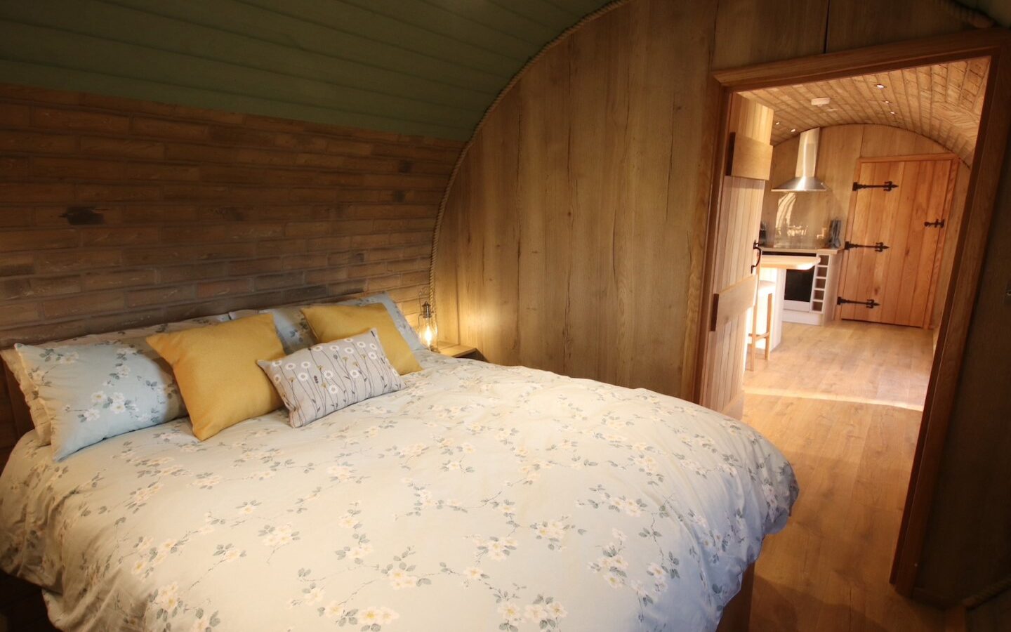 Cozy room with a double bed, floral bedding, and wooden walls. Enjoy the view into the kitchen area with its charming wooden floors, reminiscent of a snug Yorkshire burrow.