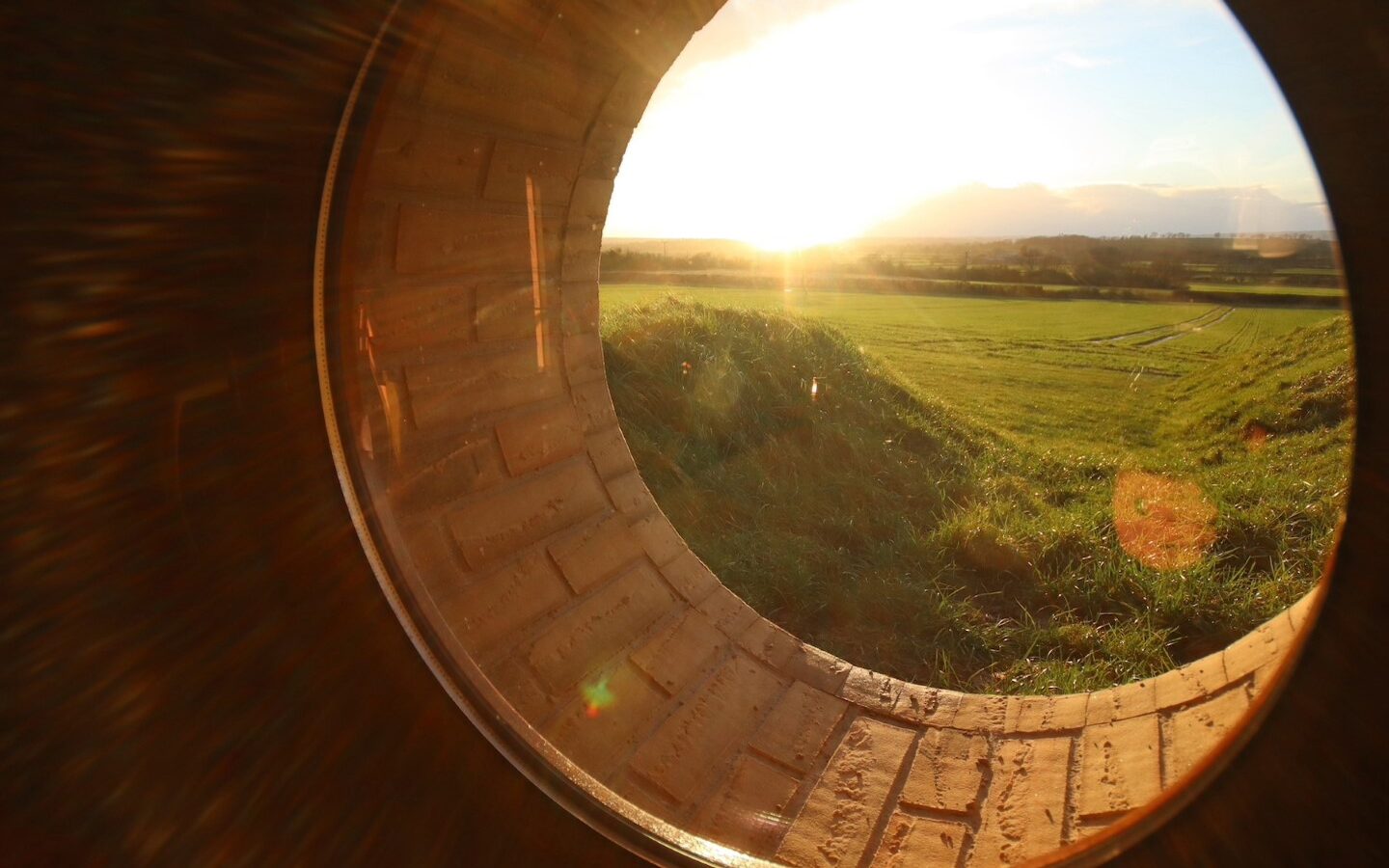 View of a grassy field and bright sunset through a circular brick tunnel opening, reminiscent of the burrows dotting the charming Yorkshire landscape.