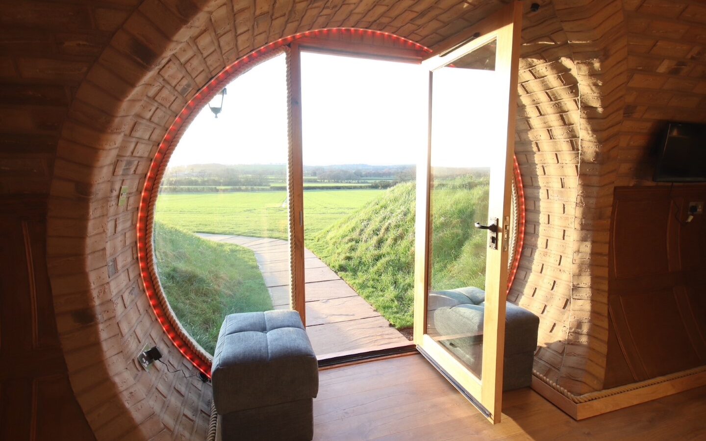 A round door with a window opens to a path leading to Yorkshire's grassy fields under a clear sky. Two small seats are nestled inside the cozy burrow.