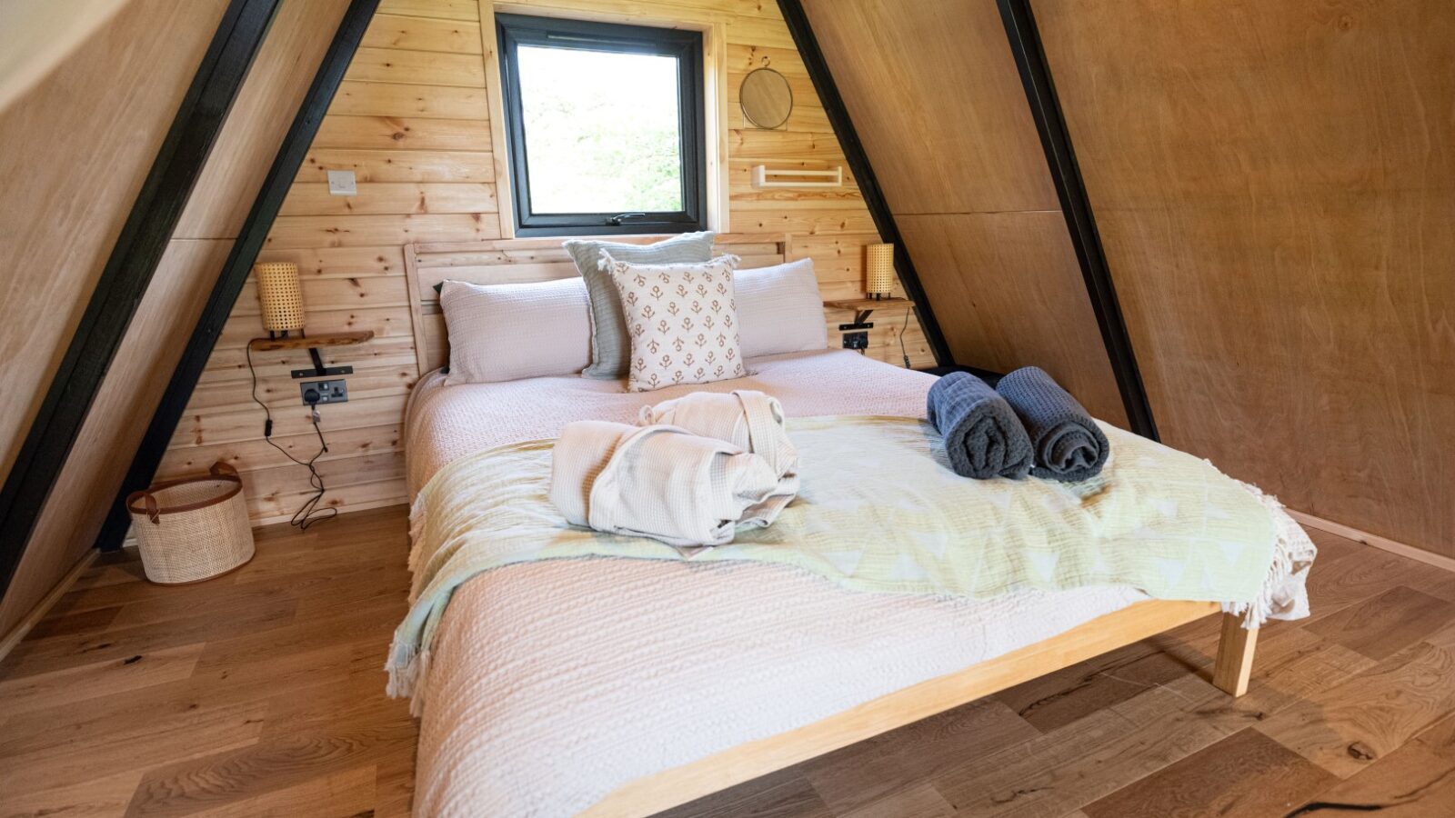 A cozy A-frame cabin bedroom at Sigrid Lodge features a wooden interior with a neatly made bed adorned with beige and patterned pillows. Two rolled towels, one gray and one blue, sit at the foot of the bed. The room has a small window above the bed, hardwood floors, and subtle decorations.