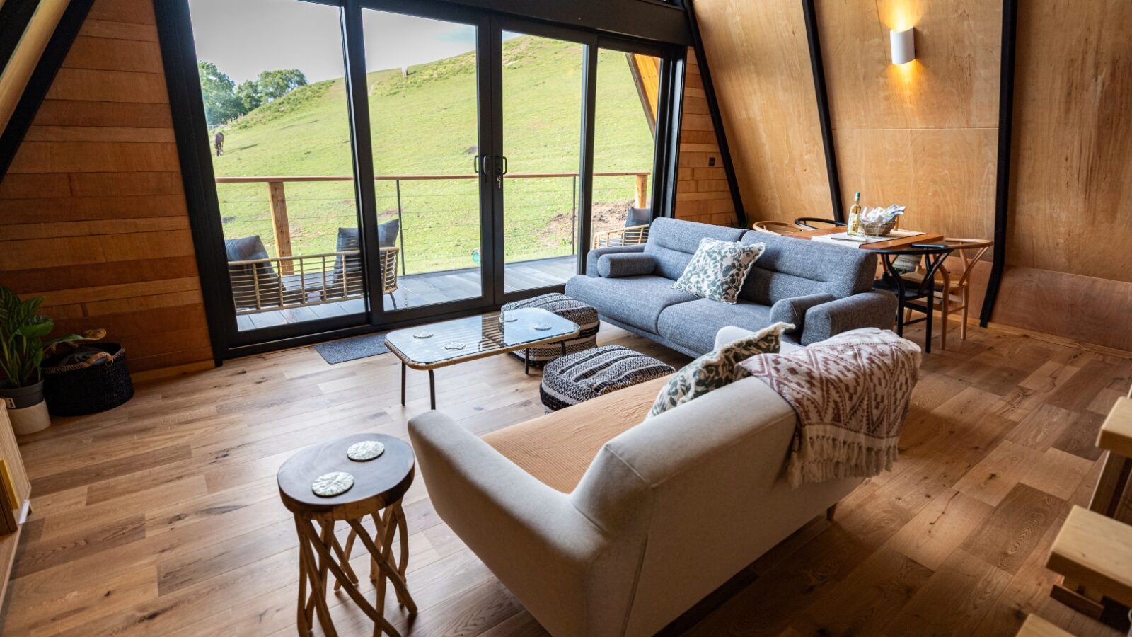 A cozy living room at Sigrid Lodge with wooden floors and walls features a grey couch and a light beige sofa. The room has a coffee table, side tables, and contemporary lighting. Large glass doors open to a deck with outdoor seating overlooking a grassy hillside.