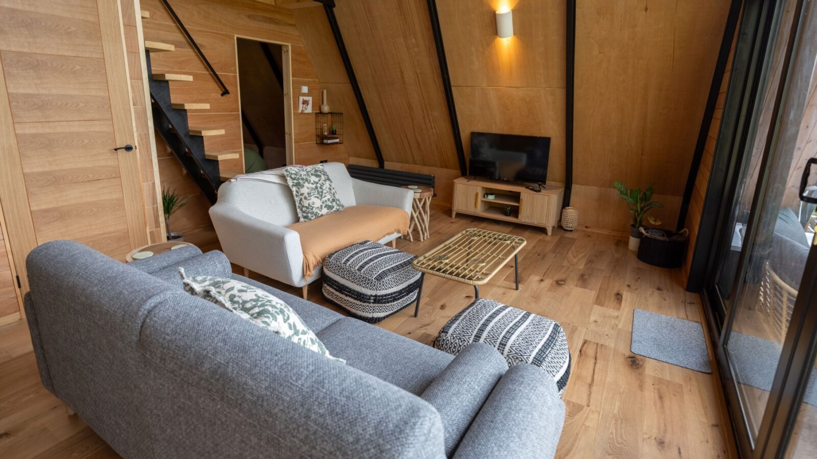 A cozy, modern living area in Sigrid Lodge boasts wooden walls and floors. The space features two sofas with patterned cushions, a TV on a stand, a small coffee table, a pouf, and potted plants. Stairs lead up to a loft, and large windows offer plenty of natural light.