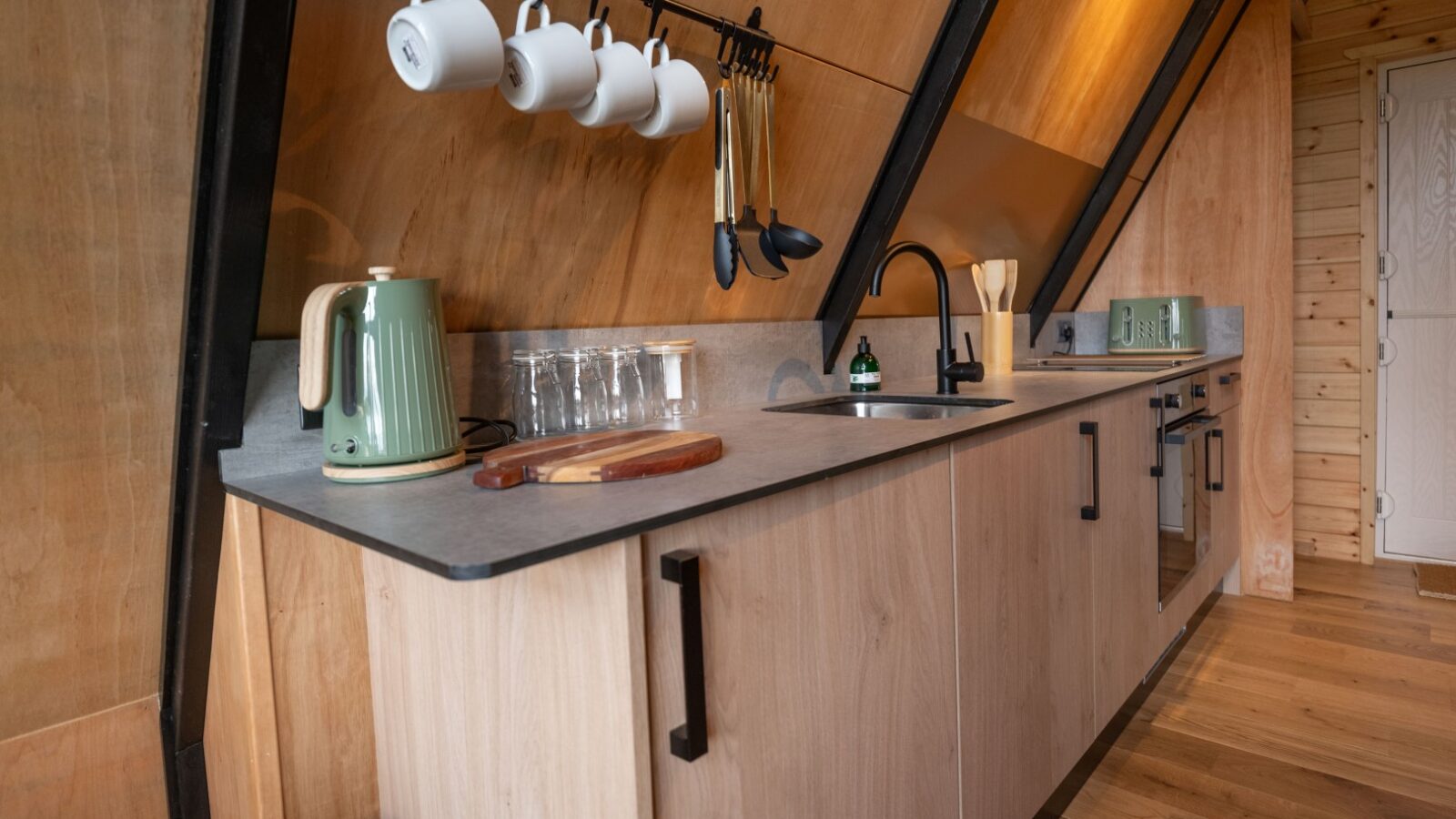 A minimalist kitchen in Sigrid Lodge with a slanted wooden ceiling features light wood cabinets, a gray countertop with a green electric kettle, a cutting board, and glassware. Mugs and utensils hang above the counter. An undermount sink and black faucet are centrally located.