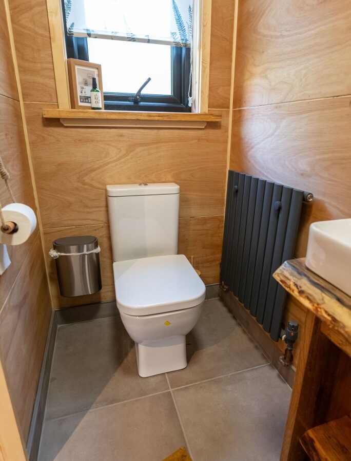 The small bathroom in Sigrid Lodge features wooden walls, a white toilet, a silver trash bin on the left, a heater on the right wall, and a wooden countertop with a white sink. A window with a floral curtain above the toilet lets in natural light.