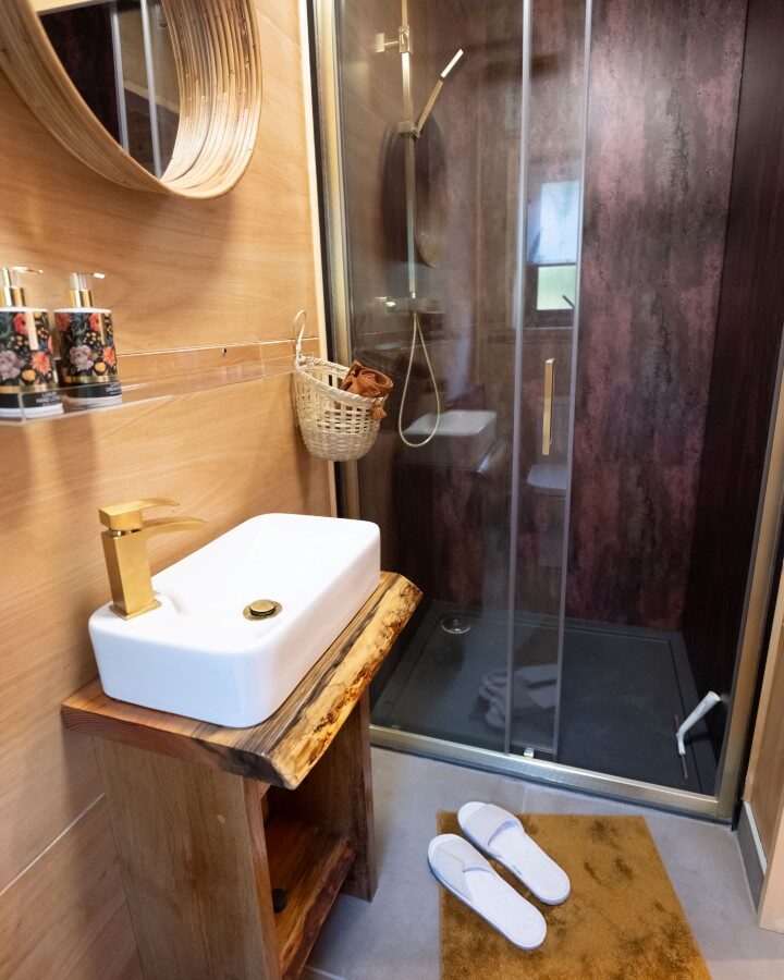 A cozy bathroom in the Sigrid Lodge style features a glass-enclosed shower with dark walls, a white sink with a wooden base, a wall-mounted round mirror, and decorative toothbrush holders. A basket is hanging on the wall, white slippers rest on a brown mat, and a towel rack is visible.