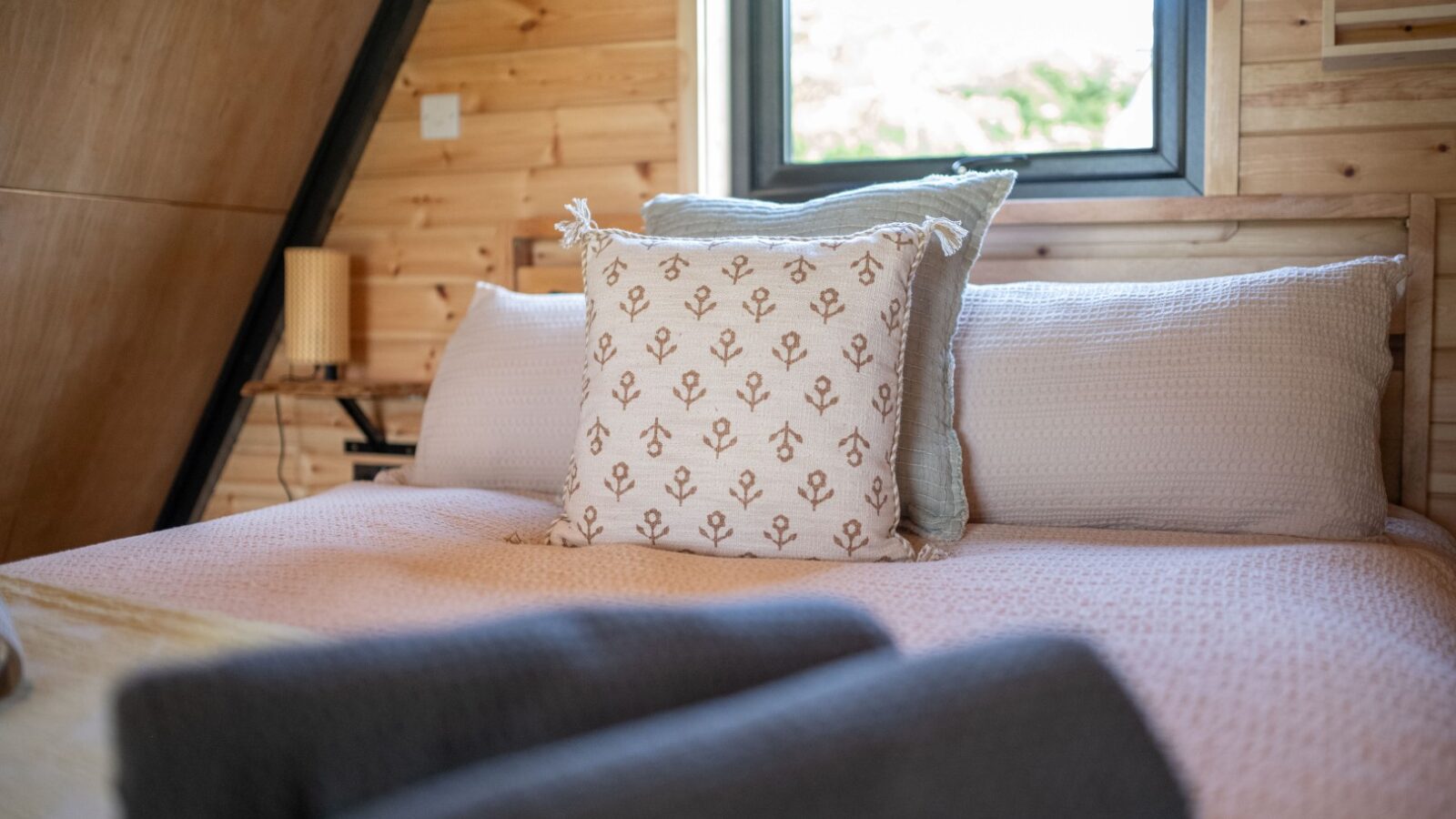 A cozy bedroom at Sigrid Lodge showcases wooden walls and a neatly made bed adorned with a decorative pillow featuring a plant pattern, along with plain pillows and cushions. A window above the bed lets in natural light, illuminating two rolled-up towels in the foreground.